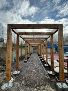 The image shows an oak framed pergola where a series of oak beams and oak columns have been erected to create the structure. The columns are set into concrete footings with stainless steel post bases, providing stability.